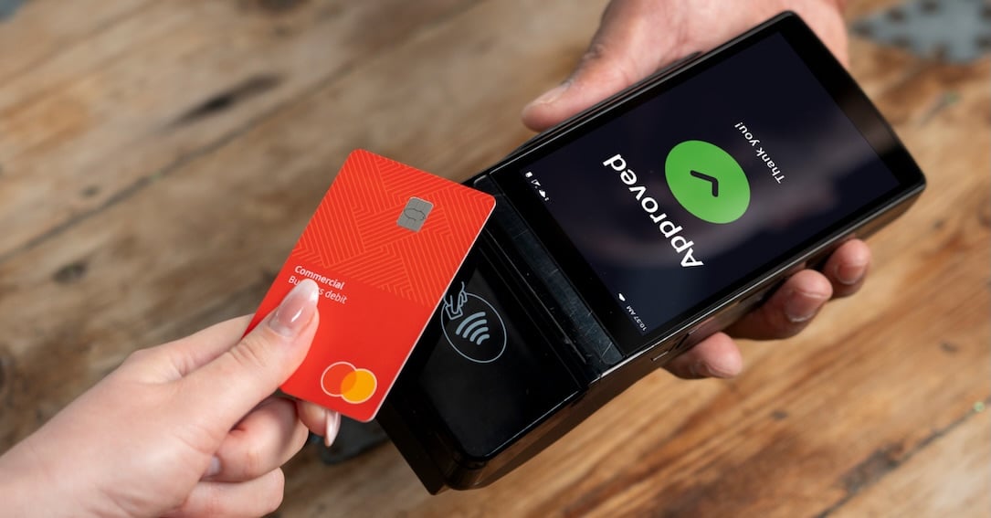 A hand holds a red Mastercard near a payment terminal displaying an "Approved" message on its screen.