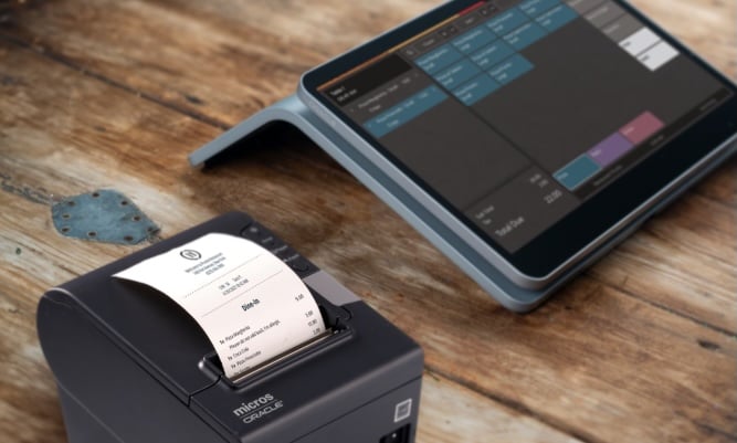 A receipt printer prints a receipt next to a tablet displaying a point-of-sale interface on a wooden table.