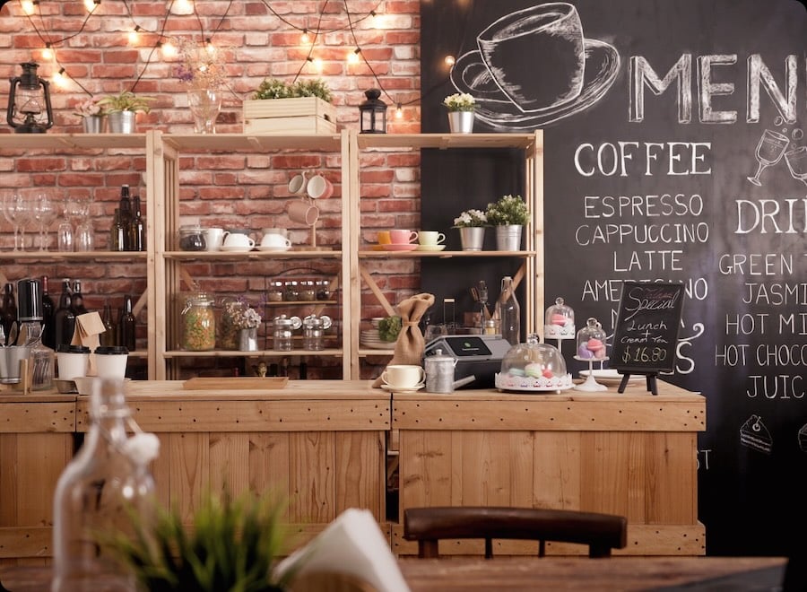 A cozy coffee shop counter with a rustic wooden design, chalkboard menu, various coffee equipment, and pastries on display. The shelves behind feature plants, mugs, and other decorative items.