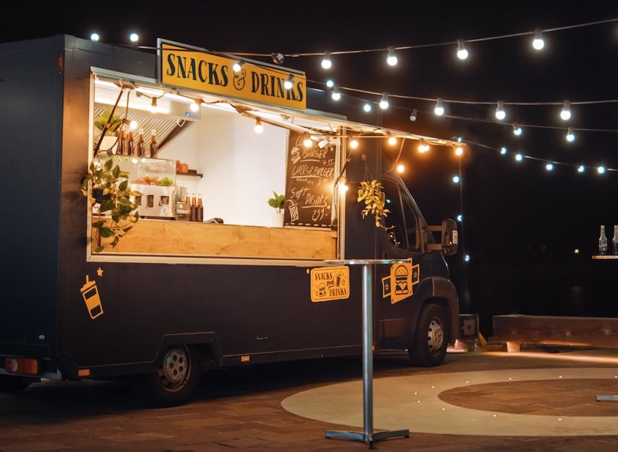 A dark-colored food truck labeled "Snacks & Drinks" is decorated with string lights and parked at a nighttime venue.