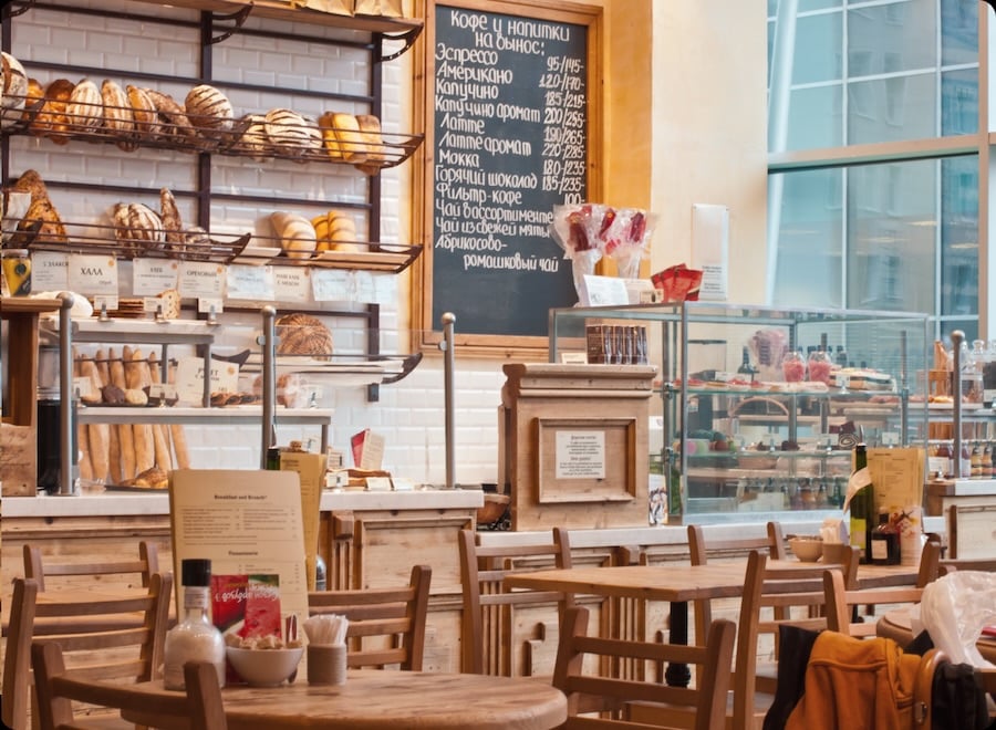 A cozy bakery with wooden tables and chairs, displaying various breads and pastries behind the counter. A chalkboard menu with prices is visible in the background.