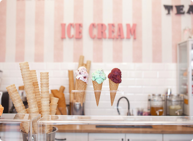 Three ice cream cones with different flavors displayed in a row in an ice cream shop with a striped pink and white background and the words "ICE CREAM" in the back.