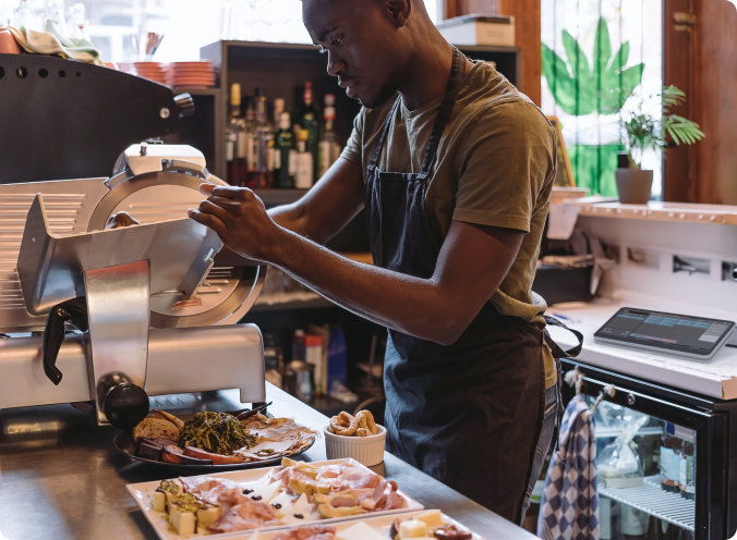 A person slices meat in a deli using a commercial meat slicer, with various sliced foods and kitchen equipment on the counter.