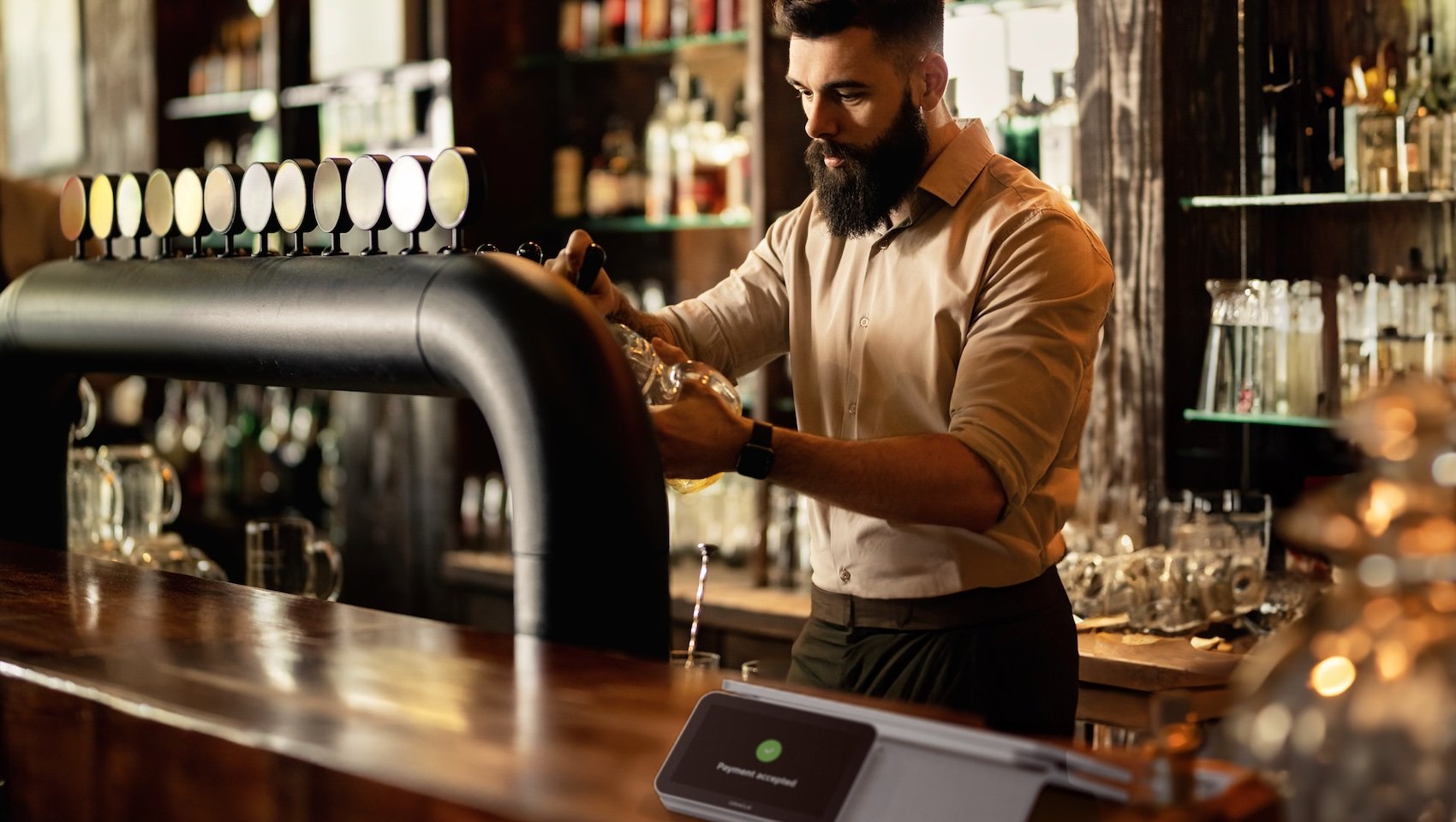 A bearded bartender in a beige shirt pours a drink from a tap in a modern bar, with bottles and glasses in the background. A tablet with a payment screen is on the counter.