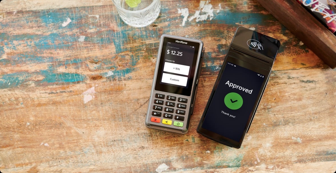 Two payment terminals sit on a wooden table. One shows a transaction of $12.25, and the other displays an "Approved" message with a green checkmark. A glass of water with a lime is in the background.