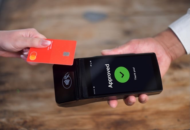 A person uses a contactless card for payment on a terminal. The terminal screen displays “Approved” with a checkmark.