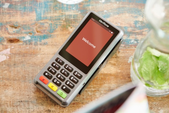A Verifone card payment terminal is placed on a wooden surface. The screen displays the word "Welcome." A glass container with mint leaves is partially visible on the right.
