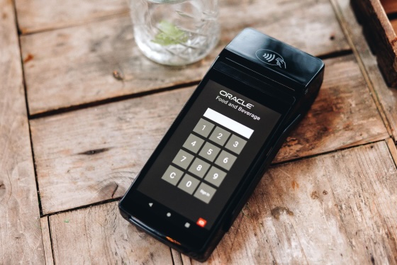 A wireless restaurant payment terminal on a wooden table displaying a numeric keypad for entering a PIN, with a glass in the background.