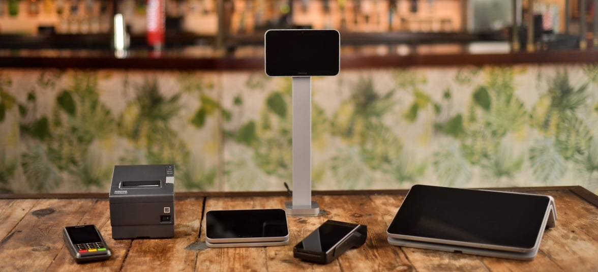 A wooden table displaying electronic devices including a smartphone, a tablet on a stand, a card reader, and other assorted gadgets. The background shows a blurred bar area with a leafy wallpaper.