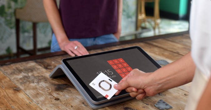 Person holds a card near a tablet with a digital keypad display at a wooden counter, while another person stands in the background.