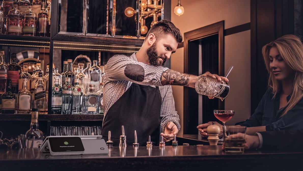 Bartender with tattoos mixing a cocktail behind the bar while a woman watches. A touchscreen payment terminal sits on the bar counter.