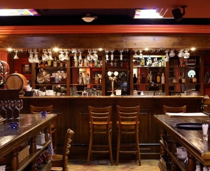 A well-lit bar with wooden chairs, tables, and fully stocked shelves displaying various bottles and glassware. The warm ambience is accentuated by stained glass accents on the ceiling.