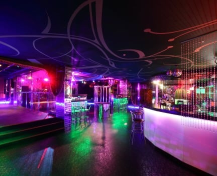 Colorful nightclub interior with neon lights, a bar counter, high stools, and a dance floor. The ceiling features artistic designs and there are reflective surfaces throughout the room.