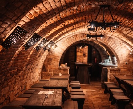 A dimly lit, rustic cellar with stone walls and arched ceilings, furnished with long wooden tables and benches, decorated with wrought iron light fixtures.