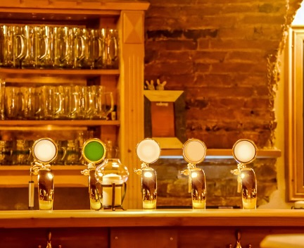 A rustic bar counter with five beer taps and empty glass mugs on shelves in the background.