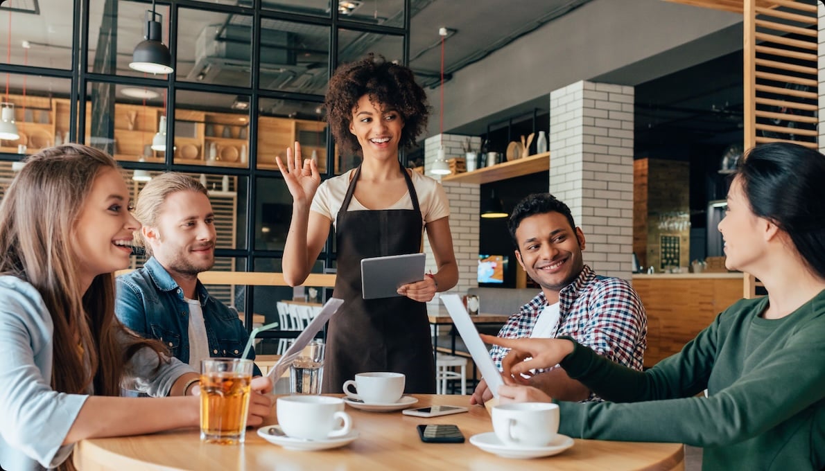 A waitress with a tablet takes orders from four seated customers at a café. The group smiles and interacts in a casual setting.