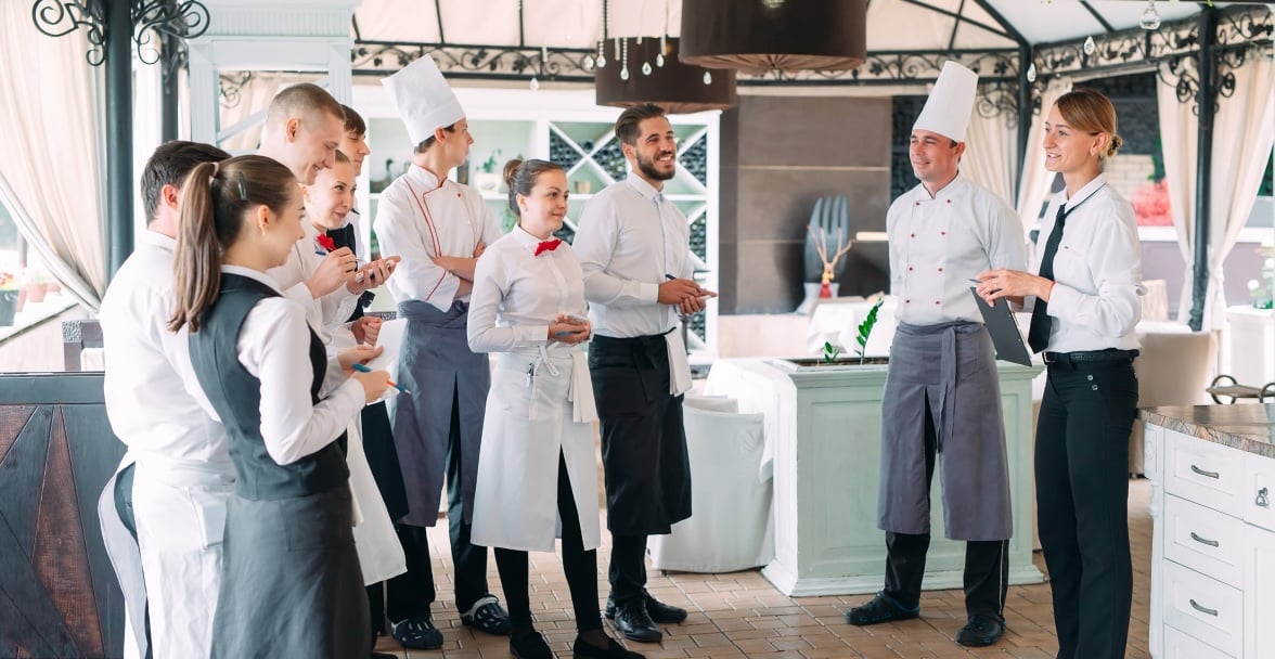 A group of restaurant staff, including chefs and servers, stands in a semi-circle, attentively listening to a female manager giving instructions.