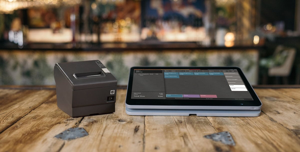 A touchscreen point-of-sale device and a receipt printer are displayed on a wooden countertop in a cozy restaurant setting.