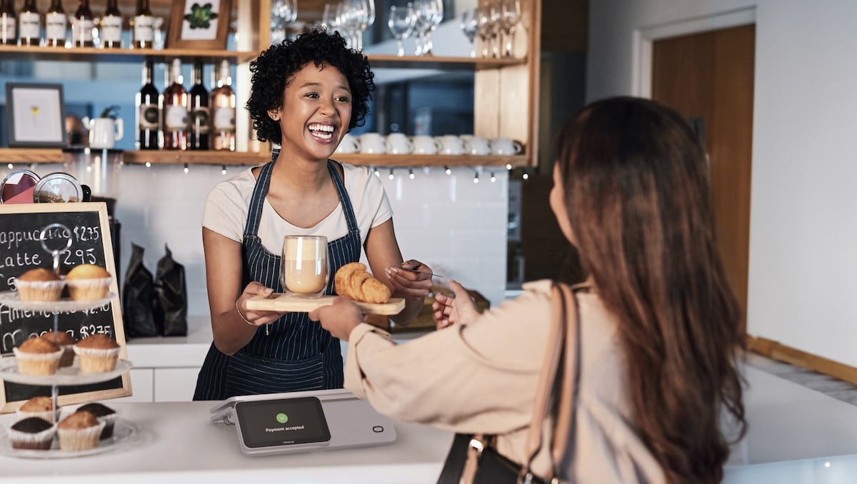 Barista in a coffee shop smiling while handing a tray with a latte and pastries to a customer across the counter.