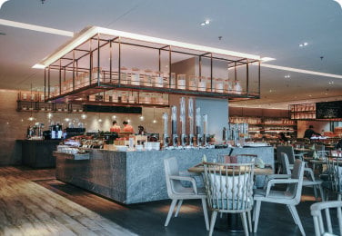 A modern restaurant interior featuring a central buffet table with food, overhead shelving, and seating areas with wooden chairs and tables. The space is well-lit with contemporary design elements.