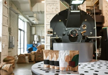 Coffee roaster with multiple bags of coffee displayed in the foreground and stacks of burlap sacks in the background.
